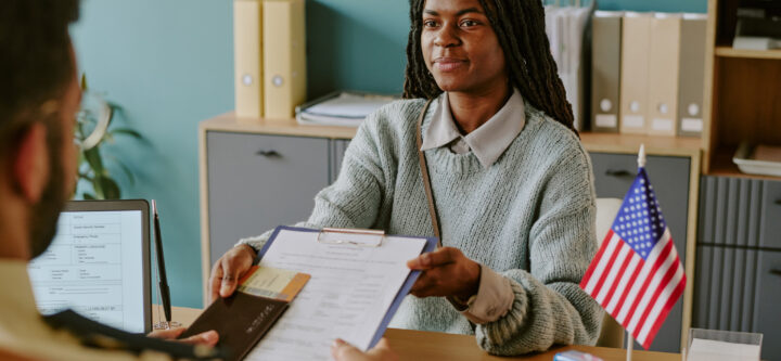 African American woman holding clipboard and pen during visa interview. Office setting with documents and American flag on desk and bookshelf in background