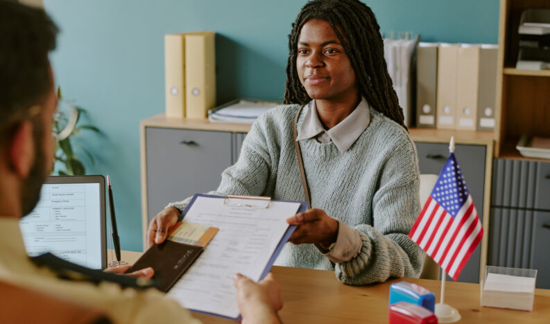 African American woman holding clipboard and pen during visa interview. Office setting with documents and American flag on desk and bookshelf in background